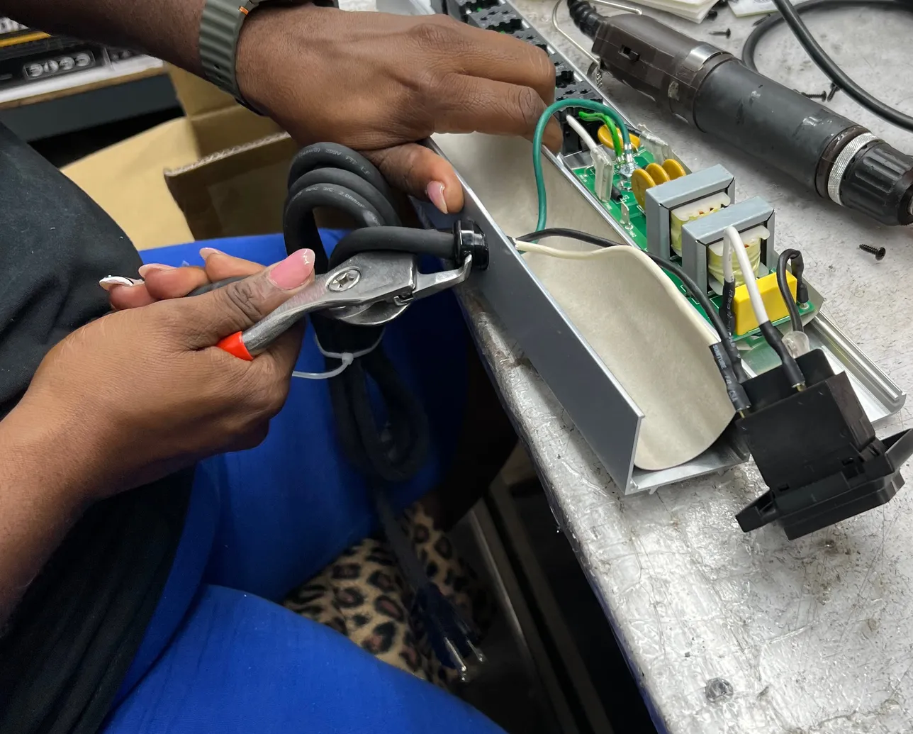 Hands using pliers to fit a power cord into the end of an open aluminum housing. Inside, a green circuit board with two transformers is wired to a black component at the far end.