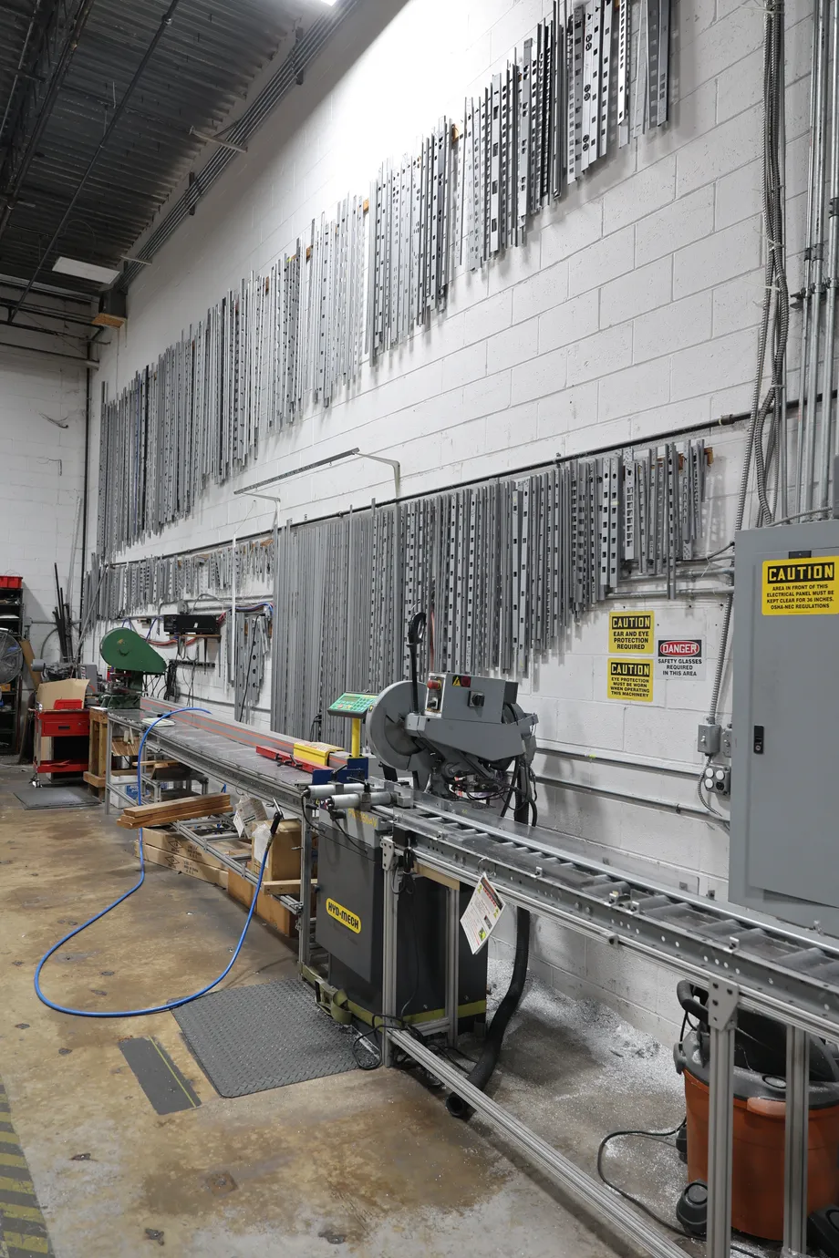 A shop wall hung with aluminum housing profiles in a range of sections and punch patterns, above the saw's roller conveyor.